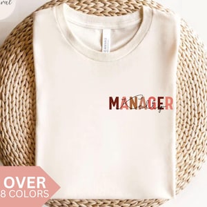 May include: A cream-coloured t-shirt featuring the word "Manager" in brown and pink lettering. The word "Kelly" is written in a cursive font below "Manager". The shirt is on a woven, circular surface. The text "Over 8 Colors" is displayed on a pink banner.
