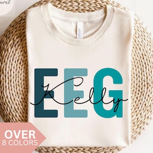 Può includere: Una maglietta bianca con il testo "EEG Kelly" in blu e turchese. Il testo è in grassetto, con un carattere a blocchi.