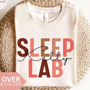 Könnte beinhalten: Cremefarbenes T-Shirt mit den Worten "SLEEP Kelly LAB" in Braun- und Rosatönen. Der Text "OVER 8 COLORS" befindet sich in einer rosa Box. Das Shirt wird auf einer gewebten, kreisförmigen Oberfläche präsentiert.