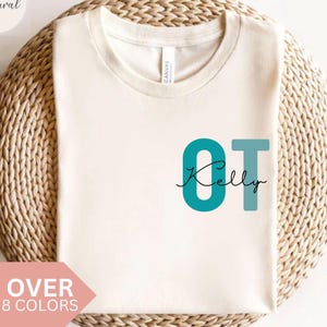 May include: Cream-colored t-shirt with the teal letters "OT" and the name "Kelly" in a script font. The shirt is displayed on a woven, circular surface. The words "Natural" and "OVER 8 COLORS" are also visible.
