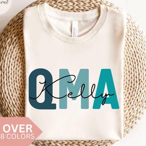 May include: Cream-colored t-shirt with the word "OMA" in teal and navy blue block letters, with "Kelly" in a cursive font. The shirt is displayed on a woven basket. Text at the bottom reads "OVER 8 COLORS".
