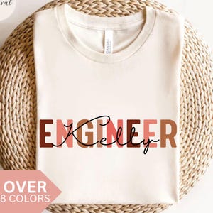 May include: Cream-colored t-shirt with the word "ENGINEER" in brown and pink letters, and the name "Kelly" in cursive. The shirt is displayed on a woven, circular surface. Text at the bottom reads "OVER 8 COLORS".