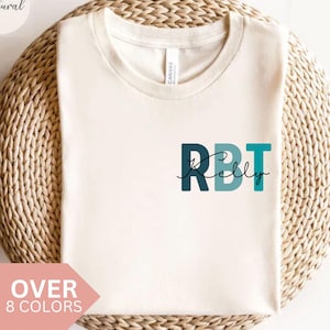 Könnte beinhalten: Cremefarbenes T-Shirt mit dem Text "RBT Kelly" in Teal und Dunkelblau. Das Shirt wird auf einer gewebten, kreisförmigen Oberfläche präsentiert. Die Worte "Over 8 Colors" stehen in einem rosa Banner.