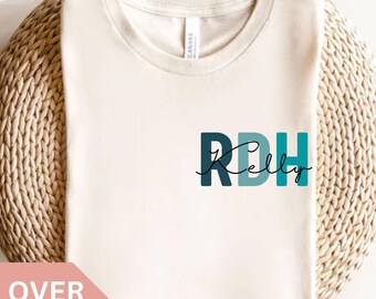 May include: Cream-coloured t-shirt with the text "RDH Kelly" in teal and dark teal. The shirt is displayed on a woven, circular surface. The words "Natural" and "OVER 8 COLORS" are also visible.