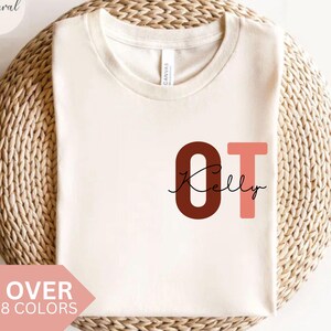 Puede incluir: Camiseta color crema con las letras "OT" en marrón y rosa, con el nombre "Kelly" en escritura cursiva. La camiseta se muestra sobre una superficie circular tejida. La pancarta rosa dice "OVER 8 COLORS".