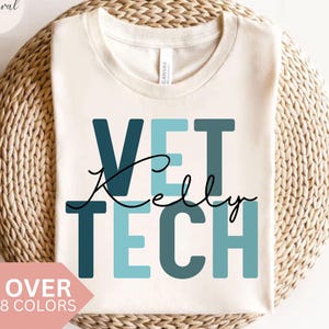 Op de afbeelding: Crèmekleurig T-shirt met de woorden "VET TECH Kelly" in teal en donkerblauwe blokletters en zwarte cursieve letters. Het shirt wordt gepresenteerd op een geweven, lichtbruin oppervlak. Een roze banner onderaan leest "OVER 8 COLORS".