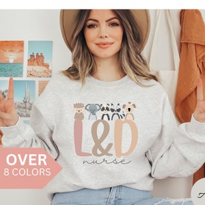 Op de afbeelding: Een lichtgrijs sweatshirt met een afbeelding van dieren rond de letters "L&D" en de tekst "nurse" eronder. Het sweatshirt is verkrijgbaar in 8 kleuren.
