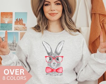 May include: A gray sweatshirt with a graphic of a bunny wearing pink-rimmed glasses and a pink bow tie. The text "OVER 8 COLORS" is visible on the bottom left of the image. The word "Ash" is visible on the bottom right of the image.