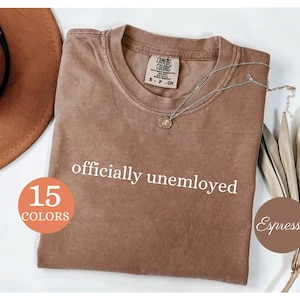 May include: A brown t-shirt with the words "officially unemployed" in white. The shirt has a crew neck and short sleeves. A silver necklace with a pendant is draped across the shirt. The image also includes a brown hat and dried plants.