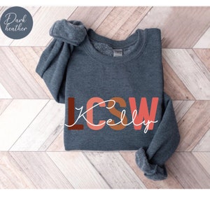 K&ouml;nnte beinhalten: Dunkelgrauer Sweatshirt mit dem Text "LCSW Kelly" in bunten Blockbuchstaben.