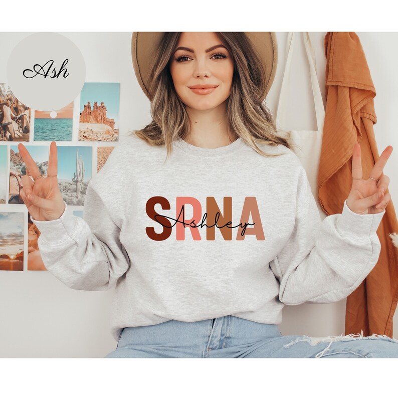May include: A gray sweatshirt with the text "SRNA" in large, colorful letters. The letters are in shades of brown, pink, and red. The text "Ashley" is written in cursive below the letters.
