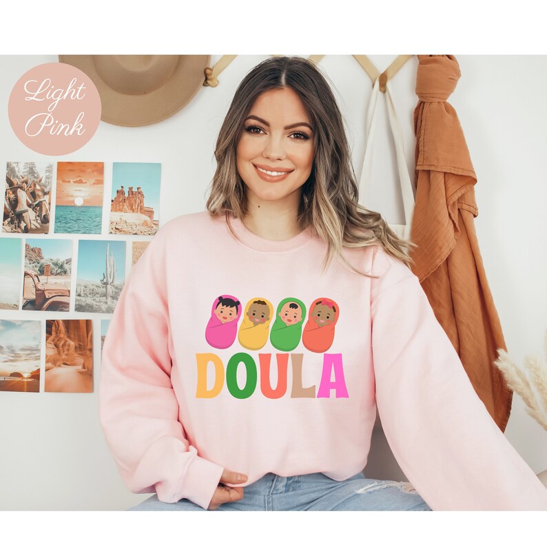 May include: A light pink sweatshirt with a graphic of four babies in swaddles and the word "DOULA" in colorful letters.