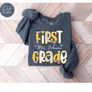 Puede incluir: Sudadera gris oscuro con las palabras "FIRST GRADE" en un dise&ntilde;o colorido de l&aacute;pices. El texto "Mrs. Johnson" est&aacute; debajo de "FIRST". La sudadera es de cuello redondo.