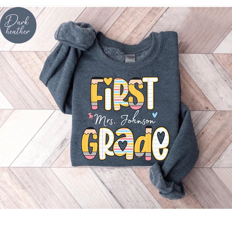 Puede incluir: Sudadera gris oscuro con las palabras "FIRST GRADE" en un dise&ntilde;o colorido de l&aacute;pices. El texto "Mrs. Johnson" est&aacute; debajo de "FIRST". La sudadera es de cuello redondo.