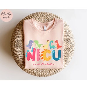 Pu&ograve; includere: Una maglietta color pesca chiaro con un design di dinosauri colorati e le parole "NICU nurse". Il design presenta vari dinosauri dei cartoni animati di diversi colori sopra la parola "NICU". Il testo "Heather peach" &egrave; nell'angolo in alto a sinistra.