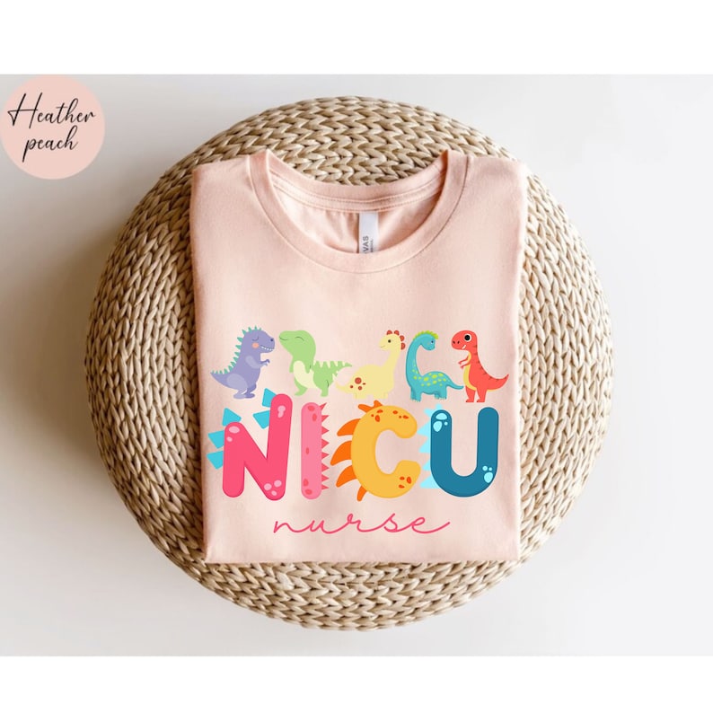 Pu&ograve; includere: Una maglietta color pesca chiaro con un design di dinosauri colorati e le parole "NICU nurse". Il design presenta vari dinosauri dei cartoni animati di diversi colori sopra la parola "NICU". Il testo "Heather peach" &egrave; nell'angolo in alto a sinistra.