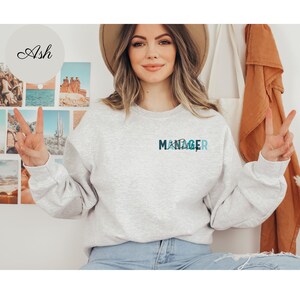 May include: A gray sweatshirt with the word "MANAGER" printed on the front in blue and teal lettering. The letters are stylized with a graphic design element.