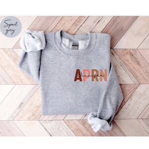 May include: A gray sweatshirt with the letters APRN in large, colorful letters. The letters are in shades of brown, pink, and orange. The word 'Jennifer' is written in cursive script below the letters APRN.