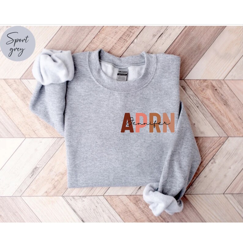 May include: A gray sweatshirt with the letters APRN in large, colorful letters. The letters are in shades of brown, pink, and orange. The word 'Jennifer' is written in cursive script below the letters APRN.