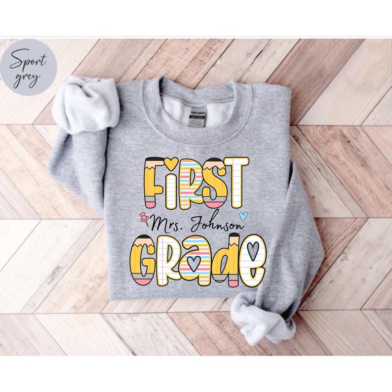 Puede incluir: Una sudadera gris claro con las palabras "FIRST GRADE" en un dise&ntilde;o colorido con tem&aacute;tica de l&aacute;pices. El texto "& Mrs. Johnson" tambi&eacute;n est&aacute; impreso en la camisa, junto con un coraz&oacute;n y un coraz&oacute;n azul. La sudadera est&aacute; doblada sobre una superficie de madera clara.