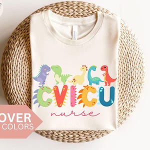 Könnte beinhalten: Cremefarbenes T-Shirt mit einem bunten Dinosaurier-Design und der Aufschrift "CVICU nurse". Die Dinosaurier sind in verschiedenen Farben gehalten, und der Text ist in einer verspielten Schriftart. Das Shirt wird auf einer gewebten Oberfläche präsentiert.
