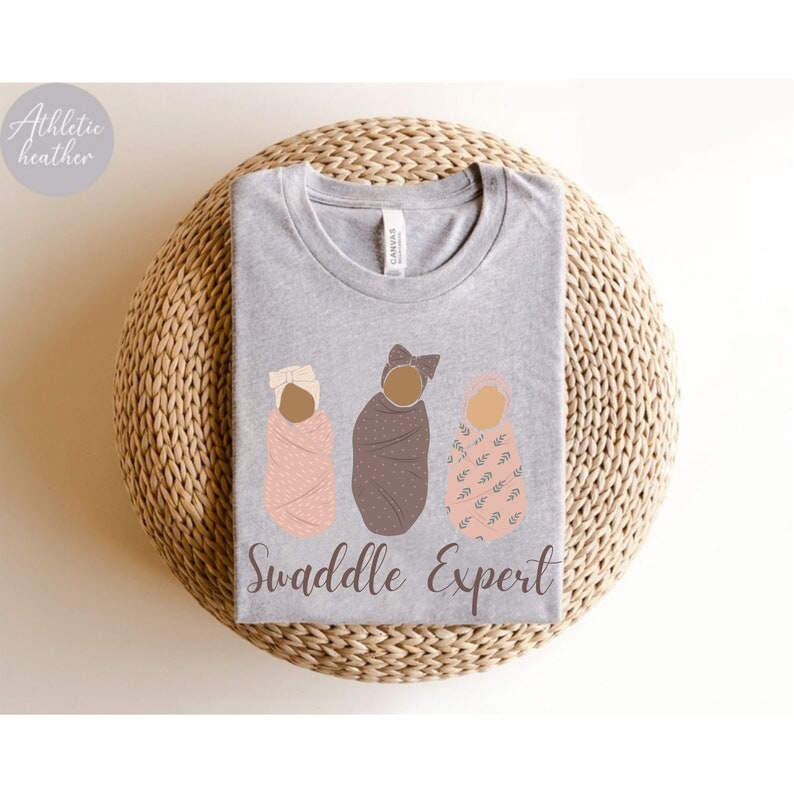 May include: A heather grey t-shirt with a graphic of three swaddled babies. The text "Swaddle Expert" is printed below the graphic.
