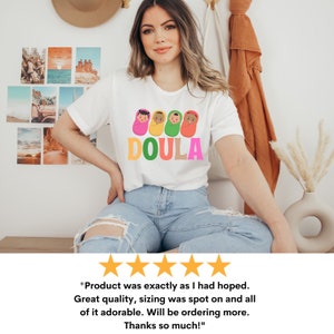May include: White t-shirt with a graphic of four colorful nesting dolls with the word "DOULA" printed below. The t-shirt is being worn by a person with long brown hair and is sitting down.