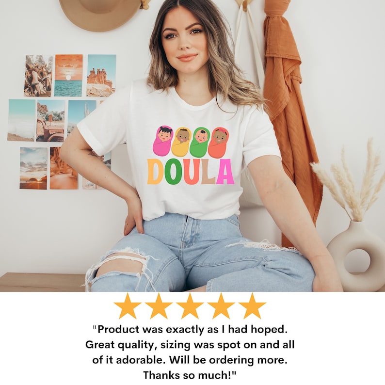May include: White t-shirt with a graphic of four colorful nesting dolls with the word "DOULA" printed below. The t-shirt is being worn by a person with long brown hair and is sitting down.