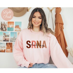 May include: A light pink sweatshirt with the letters "SRNA" in brown, pink, and orange, with the name "Ashley" written in black cursive underneath.