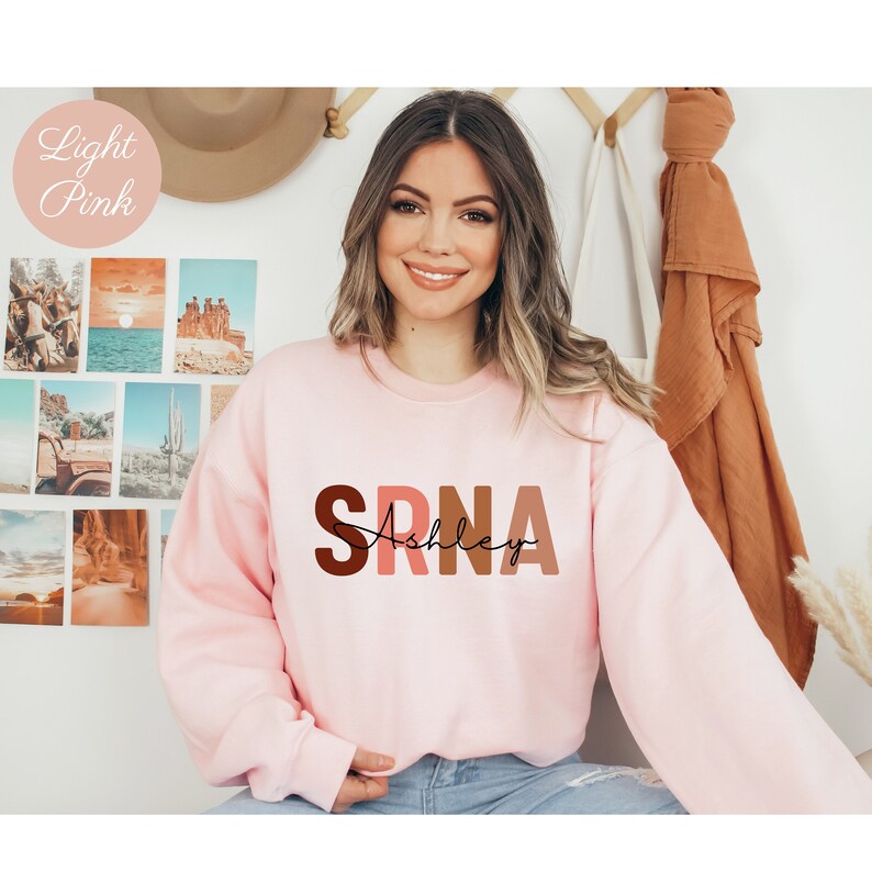 May include: A light pink sweatshirt with the letters "SRNA" in brown, pink, and orange, with the name "Ashley" written in black cursive underneath.