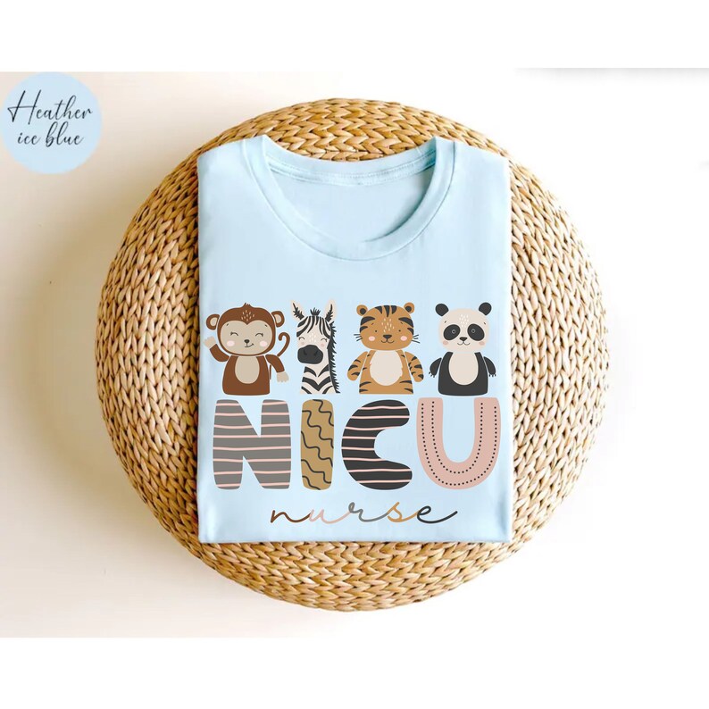 Pode incluir: T-shirt azul claro com gola redonda. A camisa apresenta um desenho com um macaco, uma zebra, um tigre e um panda acima das letras "NICU" e da palavra "nurse". A camisa &eacute; feita de um material macio.
