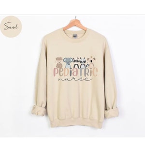 Puede incluir: Una sudadera de color arena con un gr&aacute;fico de cinco animales de dibujos animados y el texto "Pediatric Nurse".