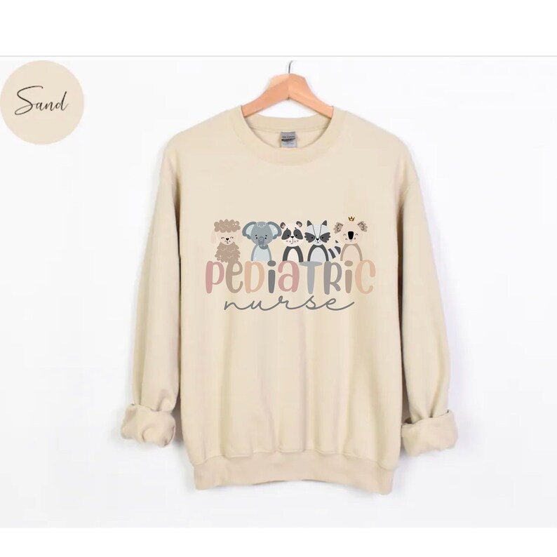 Puede incluir: Una sudadera de color arena con un gr&aacute;fico de cinco animales de dibujos animados y el texto "Pediatric Nurse".