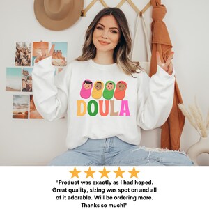 May include: White sweatshirt with a graphic of four babies in swaddles and the word "DOULA" in colorful letters. The sweatshirt is being worn by a person with long brown hair and a light complexion.