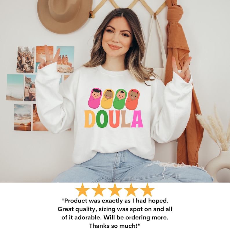 May include: White sweatshirt with a graphic of four babies in swaddles and the word "DOULA" in colorful letters. The sweatshirt is being worn by a person with long brown hair and a light complexion.