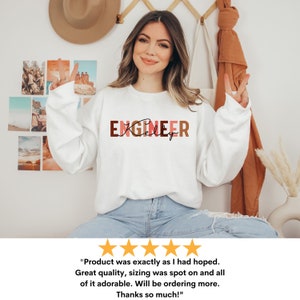 Może przedstawiać: Biała bluza z napisem "ENGINEER" w brązowych, r&oacute;żowych i czerwonych literach. Słowo "Kelly" jest napisane kursywą pod słowem "ENGINEER".