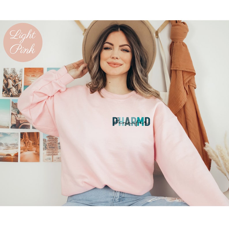 May include: A light pink sweatshirt with the word "PHARMD" printed on the front in a blue and green gradient. The sweatshirt is being worn by a person with long brown hair and a brown hat.