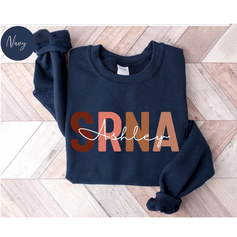 May include: Navy blue sweatshirt with the letters "SRNA" in brown, pink, and orange. The name "Ashley" is written in white cursive underneath the letters.
