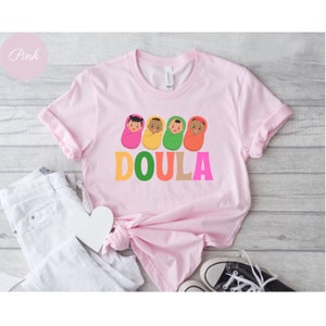May include: Pink t-shirt with a graphic of four babies wrapped in blankets and the word "DOULA" in colorful letters.
