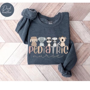 Puede incluir: Sudadera gris oscuro jaspeada con un gr&aacute;fico de cinco animales de dibujos animados y el texto "Pediatric Nurse".