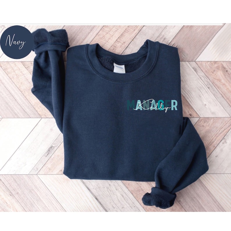 May include: Navy blue sweatshirt with the text "MANAGER" in a colorful gradient design. The text is stylized with a handwritten font.