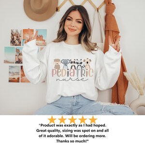 Puede incluir: Sudadera blanca con un gr&aacute;fico de una l&iacute;nea de animales de dibujos animados y el texto "pediatric nurse" en letras rosa, gris y marr&oacute;n.