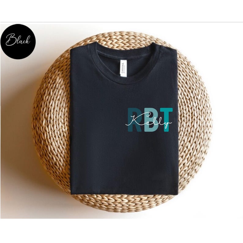 May include: Black t-shirt with the word "Kelly" in a cursive font, and the letters "RBT" in a teal and blue color scheme. The t-shirt is displayed on a woven, circular surface. A black circle with the word "Black" is in the upper left corner.