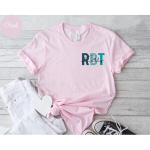 May include: A light pink t-shirt with rolled sleeves. The shirt features the text "RBT Kelly" in teal and dark teal. A white heart and white jeans are next to the shirt. The word "Pink" is in a pink circle in the upper left corner.