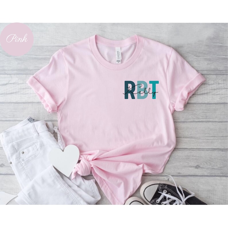 May include: A light pink t-shirt with rolled sleeves. The shirt features the text "RBT Kelly" in teal and dark teal. A white heart and white jeans are next to the shirt. The word "Pink" is in a pink circle in the upper left corner.