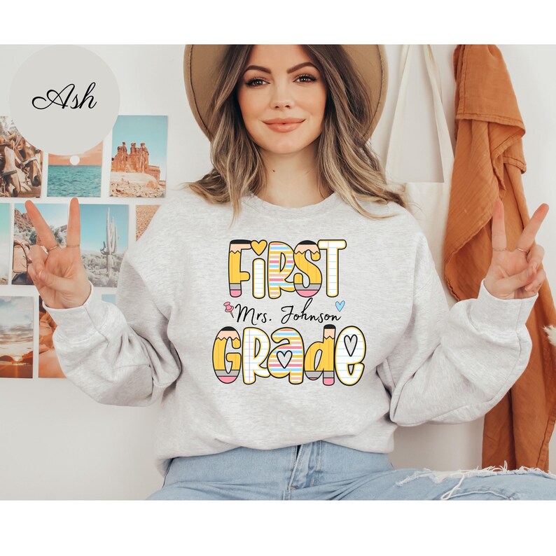 Puede incluir: Una sudadera gris claro con las palabras "FIRST Mrs. Johnson GRADE" en un dise&ntilde;o colorido con tem&aacute;tica de l&aacute;pices. La sudadera tiene mangas largas y cuello redondo. La persona de la imagen lleva vaqueros azules.