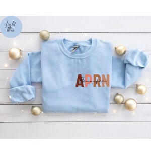 May include: A light blue sweatshirt with the letters APRN in brown, pink, and orange. The letters are stacked on top of each other. The word 'Jennifer' is written in black cursive script below the letters.