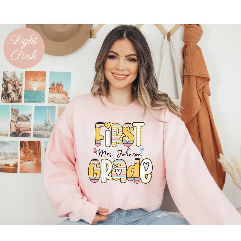 Puede incluir: Sudadera rosa claro con las palabras "FIRST GRADE Mrs. Johnson" en una fuente colorida y juguetona. El dise&ntilde;o incluye l&aacute;pices y gr&aacute;ficos de coraz&oacute;n. La sudadera es de cuello redondo y manga larga.
