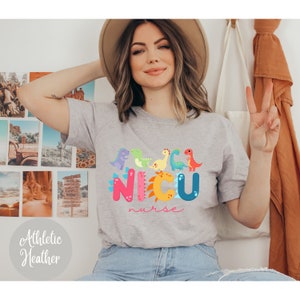 Pu&ograve; includere: T-shirt grigio atletico con un design di dinosauri colorati e la scritta "NICU nurse". Il design presenta vari dinosauri dei cartoni animati in tonalit&agrave; viola, verde, giallo, blu e rosso. Il testo "NICU" &egrave; in grandi lettere colorate.