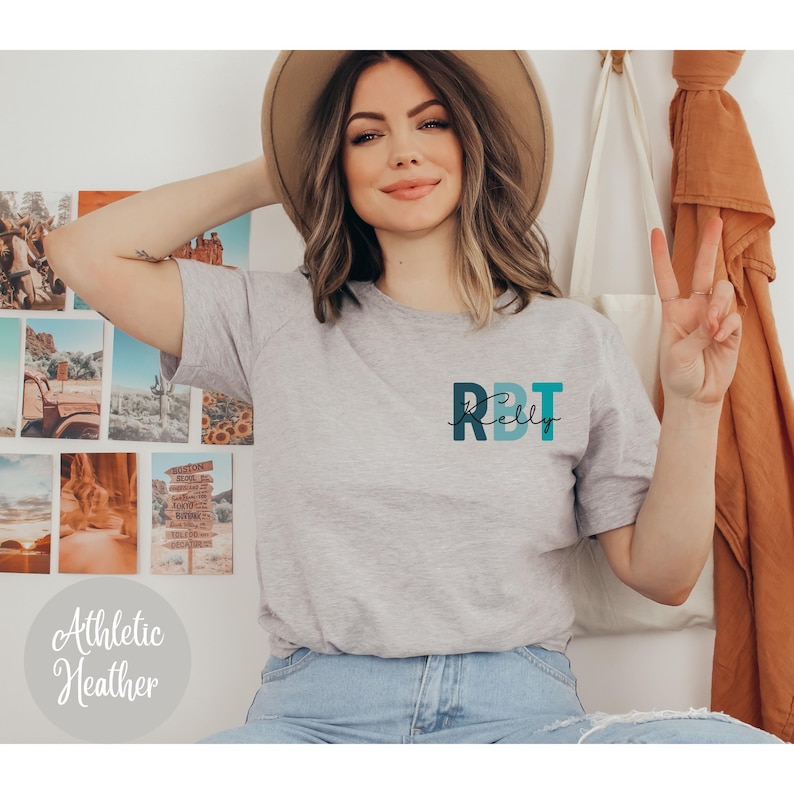 May include: Athletic heather gray t-shirt with the text "RBT Kelly" in teal and navy blue. The shirt is a short-sleeved crew neck style.